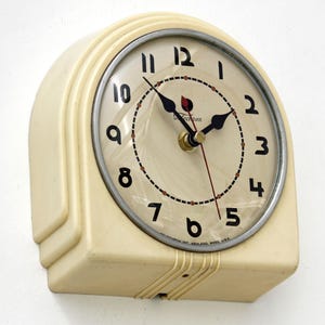 May include: An off-white Telechron wall clock with a rounded top and stepped base. The cream-coloured face features black Arabic numerals, a circular minute track with red accents, black hour and minute hands, and a thin red second hand.