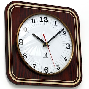 May include: Square wall clock with a dark wood grain frame and a gold-coloured inner rim. The clock face is white with black numerals and hands, and a red second hand. The clock reads "JAZ Transistor" and "France".