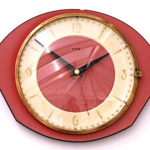 May include: A wall clock with a red, textured, eye-shaped frame and a circular cream-coloured face. The clock features black hour and minute hands, a thin red second hand, and the text FFR printed in black. It has a gold-toned metal bezel.