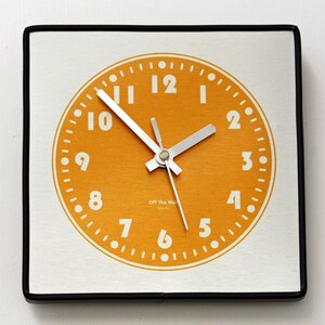 May include: Square wall clock with a brushed silver face and a black frame. The clock face is orange with white numerals and hands. The text "Off The Wall Clocks" is printed at the bottom of the clock face.