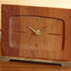 May include: A rectangular wooden clock with a light brown finish. The clock face is a lighter shade of brown with gold-coloured hands and markers. The clock has a gold-coloured base and a rectangular frame.