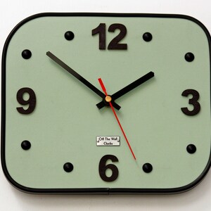 May include: A square, pale green wall clock with black numerals at 3, 6, 9, and 12. The clock has a black border, black dots for hour markers, and black hour and minute hands. The second hand is red. The clock face has the text "Off The Wall Clocks".