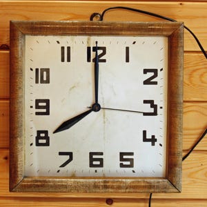 May include: Square wall clock with a light-coloured face and black numerals. The clock has a wooden frame and black hands, indicating the time is approximately eight o'clock. The clock is hanging on a wooden wall.