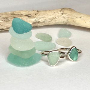 May include: Two silver rings with sea glass stones in shades of green and blue. A stack of sea glass stones and a piece of driftwood are also in the image. The rings are a unique jewelry item.