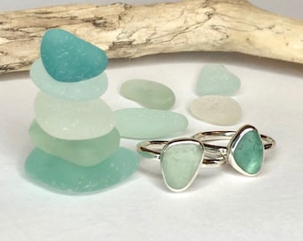 Puede incluir: Dos anillos de plata con piedras de vidrio marino en tonos verdes y azules. Una pila de piedras de vidrio marino y un trozo de madera flotante también están en la imagen. Los anillos son una joya única.