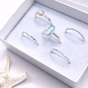 Puede incluir: Una colecci&oacute;n de anillos de plata exhibidos en una caja blanca. Los anillos presentan varios dise&ntilde;os, incluyendo una piedra redonda, un vidrio de mar ovalado y una banda de cuentas. Una estrella de mar blanca es visible, a&ntilde;adiendo un tema costero.