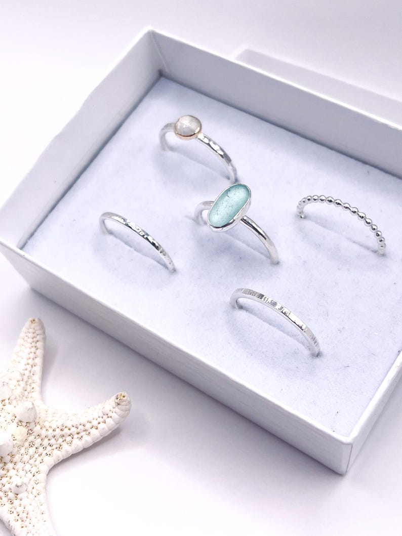 Puede incluir: Una colecci&oacute;n de anillos de plata exhibidos en una caja blanca. Los anillos presentan varios dise&ntilde;os, incluyendo una piedra redonda, un vidrio de mar ovalado y una banda de cuentas. Una estrella de mar blanca es visible, a&ntilde;adiendo un tema costero.