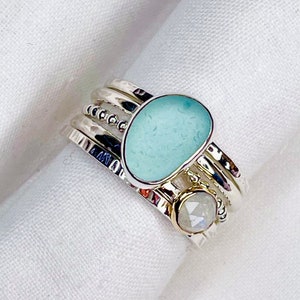 May include: A silver-toned stackable ring set. The rings feature a light blue sea glass stone and a smaller, round, iridescent stone. The bands have hammered and beaded details. The set is displayed on a white surface, showcasing the ring's textures.
