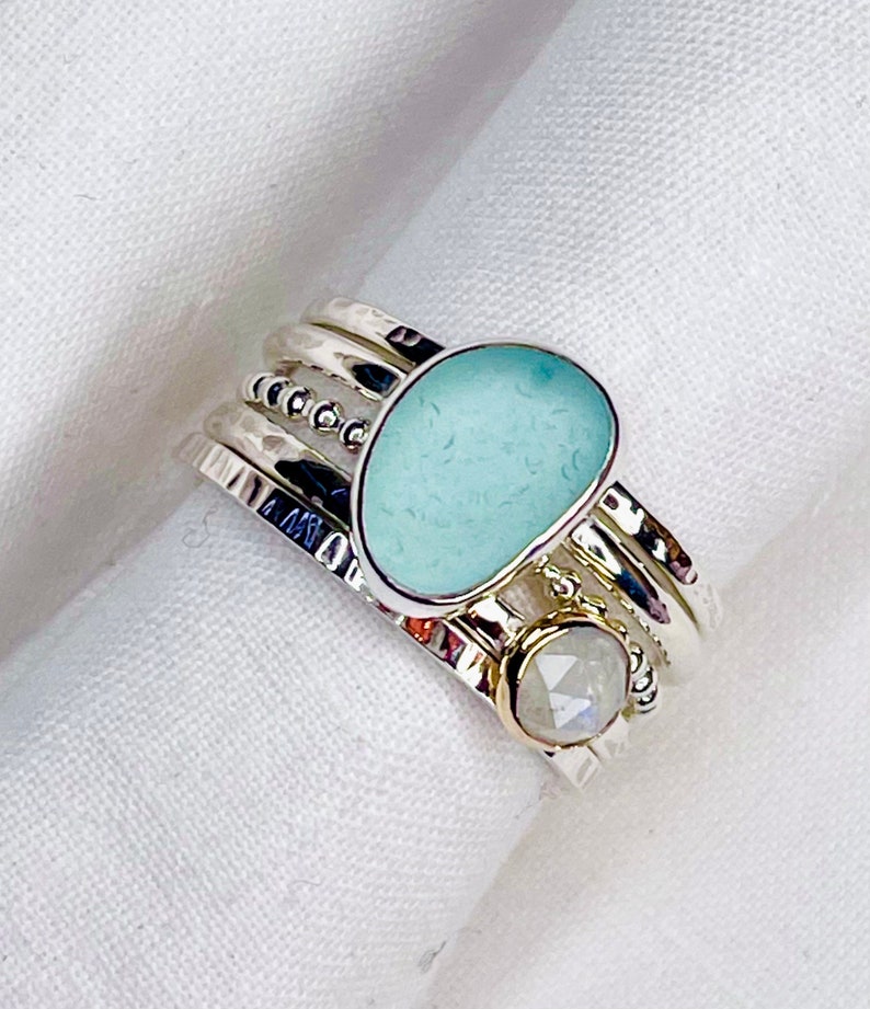 Puede incluir: Juego de anillos apilables plateados. El conjunto presenta una piedra de vidrio marino azul claro y una piedra redonda iridiscente m&aacute;s peque&ntilde;a. Los anillos tienen detalles martillados y con cuentas. Presentado sobre una superficie blanca.