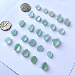 Puede incluir: Una variedad de piezas de vidrio de mar en tonos azules y verdes, dispuestas sobre una superficie blanca. Dos monedas se colocan cerca para comparar el tama&ntilde;o. Las piezas de vidrio var&iacute;an en forma y tama&ntilde;o, con una apariencia suave y esmerilada.