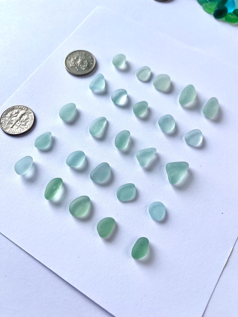 Puede incluir: Una variedad de piezas de vidrio de mar en tonos azules y verdes, dispuestas sobre una superficie blanca. Dos monedas se colocan cerca para comparar el tama&ntilde;o. Las piezas de vidrio var&iacute;an en forma y tama&ntilde;o, con una apariencia suave y esmerilada.