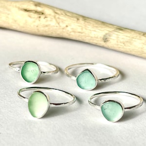 May include: Four rings featuring smooth, light green sea glass set in thin, hammered sterling silver bands. Each ring showcases a unique, rounded piece of frosted sea glass, highlighting the contrast between the polished metal and the matte, translucent green glass surfaces.