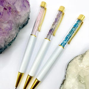 May include: Four white pens with gold detailing and crystal-filled barrels. The crystals are pink, purple, and blue. The pens are presented on a white surface with amethyst and quartz crystal formations.