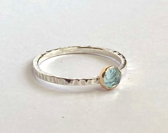 Puede incluir: Un anillo de plata con una banda texturizada y un engaste de oro que sostiene una piedra preciosa redonda de color azul claro. El anillo se encuentra sobre un fondo blanco, resaltando el diseño delicado y el color de la gema.