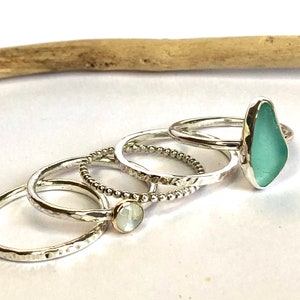 Puede incluir: Una colecci&oacute;n de anillos de plata. Un anillo presenta una piedra de vidrio marino azul claro. Otros anillos tienen bandas texturizadas, una peque&ntilde;a piedra blanca y un dise&ntilde;o de cuentas. Los anillos se exhiben sobre una superficie blanca.