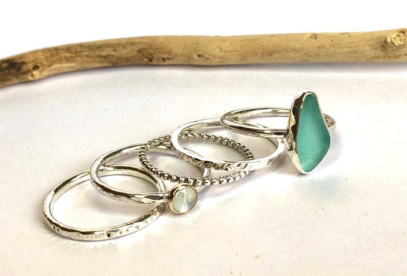 Puede incluir: Una colecci&oacute;n de anillos de plata. Un anillo presenta una piedra de vidrio marino azul claro. Otros anillos tienen bandas texturizadas, una peque&ntilde;a piedra blanca y un dise&ntilde;o de cuentas. Los anillos se exhiben sobre una superficie blanca.