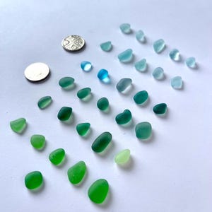 Puede incluir: Una colecci&oacute;n de trozos de vidrio de mar en tonos verdes y azules, dispuestos sobre una superficie blanca. El vidrio de mar var&iacute;a en tama&ntilde;o y forma, con algunas monedas para la escala. Los colores van desde el aguamarina claro hasta el verde esmeralda oscuro.