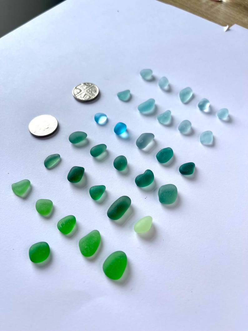 Puede incluir: Una colecci&oacute;n de trozos de vidrio de mar en tonos verdes y azules, dispuestos sobre una superficie blanca. El vidrio de mar var&iacute;a en tama&ntilde;o y forma, con algunas monedas para la escala. Los colores van desde el aguamarina claro hasta el verde esmeralda oscuro.