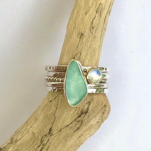 Puede incluir: Un conjunto de anillos apilables plateados. Un anillo presenta una piedra de vidrio marino verde agua, y otro una piedra lunar. Las bandas tienen detalles texturizados y con cuentas. El vidrio marino tiene una forma irregular, y la piedra lunar tiene un brillo nacarado.