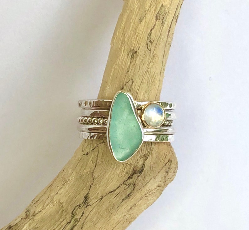 Puede incluir: Un conjunto de anillos apilables plateados. Un anillo presenta una piedra de vidrio marino verde agua, y otro una piedra lunar. Las bandas tienen detalles texturizados y con cuentas. El vidrio marino tiene una forma irregular, y la piedra lunar tiene un brillo nacarado.