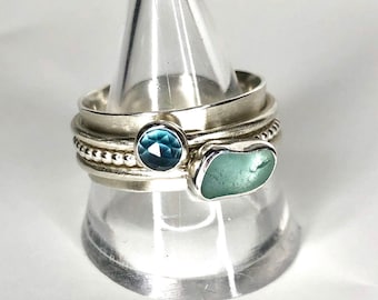 Puede incluir: Un anillo de plata con tres bandas, que presenta una piedra preciosa de topacio azul y un trozo de vidrio de mar. El anillo se muestra en un soporte de anillo transparente.