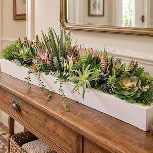 May include: A long, rectangular white planter box filled with a dense arrangement of various artificial succulents, including spiky aloe-like plants, trailing vines, and rosette-shaped foliage in shades of green, pink, and deep red.