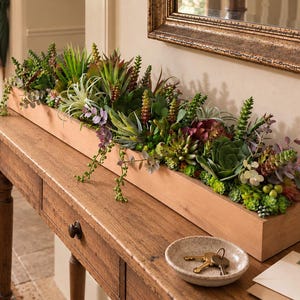 May include: A long, rectangular wooden planter box filled with a variety of artificial succulents in shades of green, purple, and red. The arrangement includes spiky, rounded, and trailing plant varieties, creating a dense, textured display.