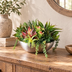 May include: A silver-toned, hammered metal bowl filled with an assortment of artificial succulents. The arrangement features various green, lime, and reddish-pink plants, including spiky agave-like leaves and trailing stems, creating a dense display.