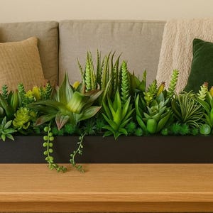Puede incluir: Una jardinera rectangular negra llena de suculentas artificiales en varios tonos de verde y amarillo. La jardinera está sobre una mesa de madera. Un sofá beige y cojines son visibles en el fondo.