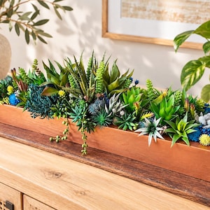 May include: A long, rectangular wooden planter box filled with a dense arrangement of various artificial succulents in shades of green, blue, and yellow. The plants feature diverse textures, including spiky, rounded, and trailing varieties.