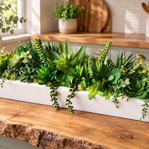 May include: A long, rectangular white wooden planter box filled with a dense arrangement of various artificial green succulents, including rosette-shaped plants, spiky varieties, and trailing string-of-pearls vines spilling over the front edge.