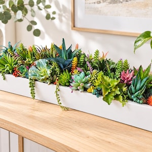 May include: A long, rectangular white planter filled with a dense arrangement of artificial succulents in various shades of green, pink, orange, and yellow. The plants feature diverse textures, including spiky, rounded, and trailing foliage.