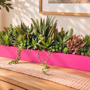 May include: A long, rectangular, bright pink wooden planter box filled with a variety of artificial succulents. The arrangement includes green, spiky aloe-like plants, trailing string of pearls, and textured, reddish-brown desert foliage.