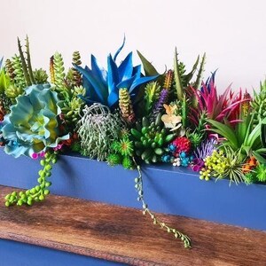 Puede incluir: Una jardinera rectangular azul llena de una variedad de suculentas artificiales coloridas. El arreglo incluye plantas verdes, azules, rosas y amarillas, creando una exhibición vibrante. La jardinera está sobre una superficie de madera.