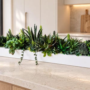 Op de afbeelding: Een lange, rechthoekige witte plantenbak gevuld met een assortiment kunstmatige groene vetplanten en cactussen. Het arrangement bevat diverse texturen, waaronder puntige bladeren, ronde rozetten en hangende stengels in de bak.