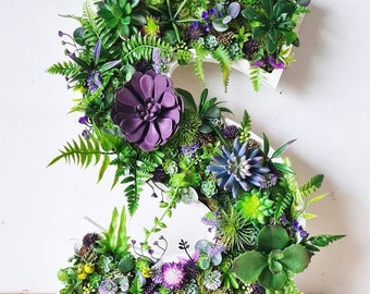 May include: A white letter S decorated with green and purple artificial succulents and plants. The letter is standing on a wooden surface.
