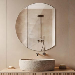 May include: A round, stone sink with a rough texture sits on a wooden countertop. A round mirror with a black frame hangs above the sink. The mirror reflects a shower with a black shower head and black fixtures.