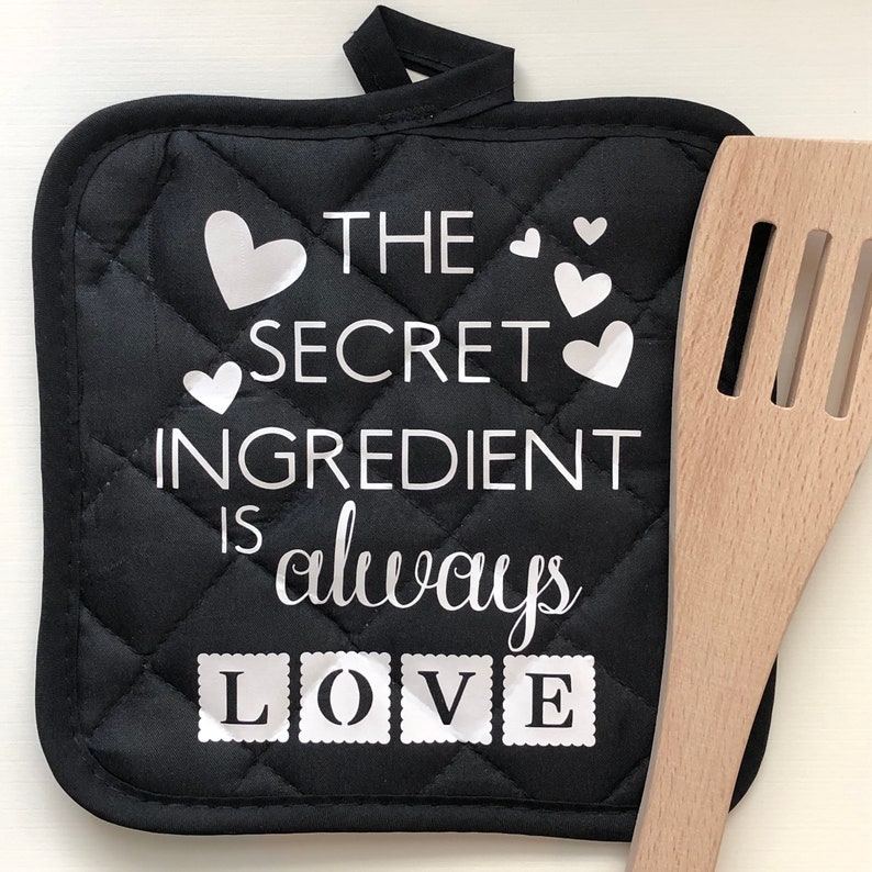 May include: Black quilted oven mitt with white text that reads "The Secret Ingredient is always LOVE" with hearts and block letters. A wooden spatula is next to the mitt.