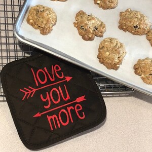 May include: A brown quilted hot pad with the text "love you more" in red lettering with arrows pointing to the text.