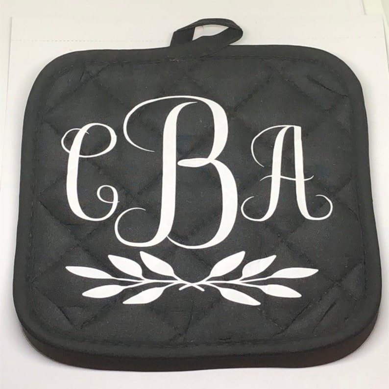 May include: Black quilted pot holder with a white monogram "CBA" and a white leafy design.