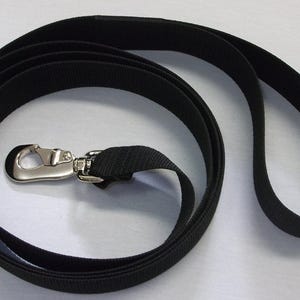 May include: A black dog leash with a silver-colored metal clasp. The leash is made of a durable, woven material and has a loop handle. The leash is coiled on a white surface.