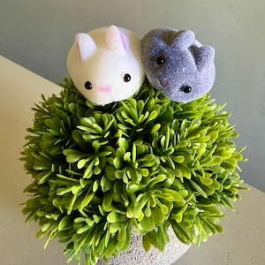 May include: A decorative arrangement featuring two plush bunny figures atop a vibrant green artificial plant. One bunny is white with pink accents, the other is gray. The plant is in a textured, light-colored pot.