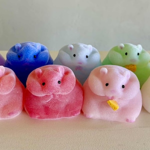 May include: A collection of colorful, hamster-shaped figurines in shades of purple, blue, white, green, yellow, and pink. Some hold small, yellow, cheese-shaped objects. The figurines have small black eyes and pink noses.
