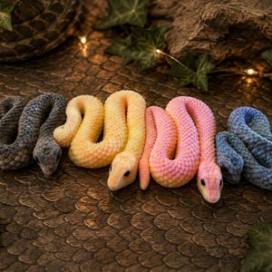 May include: Four plush snake toys arranged in a row, featuring textured fabric scales. The snakes are colored in dark gray, pale yellow, light pink, and blue. Each snake has a distinct head shape with dark eyes and a coiled, flexible body design.
