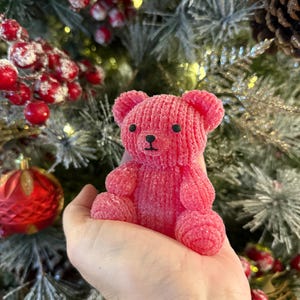 May include: A small, pink teddy bear with a textured, knitted appearance. The bear has black eyes and a black nose. The bear is held in a hand, with a Christmas tree and ornaments in the background.