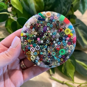 May include: A round, clear resin coaster filled with colorful beads, sequins, and small letter beads spelling out "CMF". The coaster has a light pink and yellow background and is held by a hand.