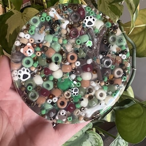 May include: A round, clear resin coaster with a variety of colorful beads, small animal figurines, and paw prints embedded in it. The coaster has a brown, green, and white color scheme.