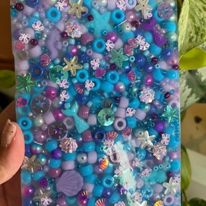 May include: A rectangular resin tray filled with a dense arrangement of blue, purple, and lavender beads. The surface features embedded charms including silver starfish, iridescent seashells, and purple mermaid tails, creating a textured, aquatic-themed mosaic.