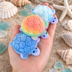 May include: Three small turtle-shaped figures with a fuzzy, textured surface. The bottom turtle is solid blue, the middle turtle features a gradient of orange, yellow, and green, and the top turtle is light blue. Each has a hexagonal shell pattern.