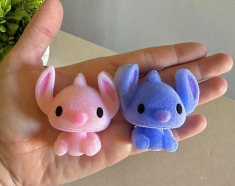 May include: Two small, flocked figurines shaped like creatures with large ears and round black eyes. One figurine is light pink, and the other is light blue. Both have a soft, fuzzy texture and are depicted in a seated position with small limbs.